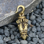 A piece of Kaya Movement's Ear Weights Blind Justice, crafted from brass, lies on a bed of small gray pebbles. The hook features two intricately carved elephant figures facing each other, ending in a pointed tip and adorned with detailed patterns and textures. This elegant item resembles traditional tribal ear hangers or piercing body jewelry.