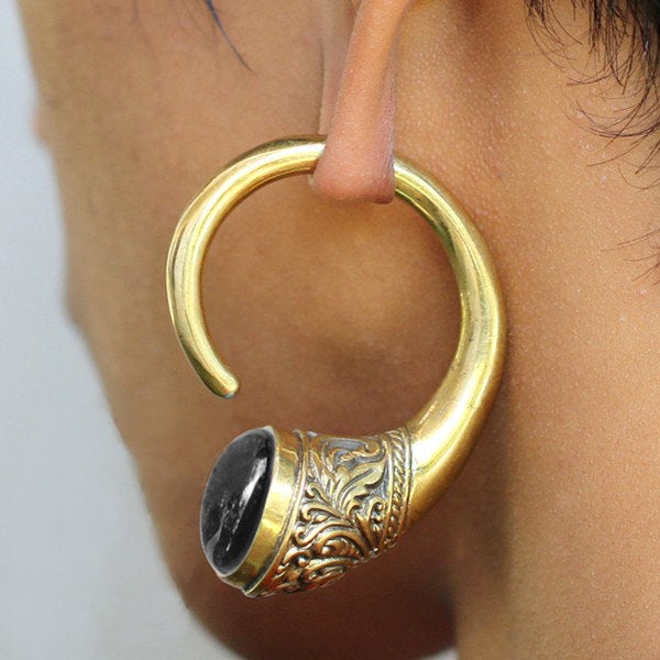 Close-up of an ear adorned with the IRO Dangle Ear Weights in Gold & Golden Obsidian by Kaya Movement, shaped like a spiral and featuring intricate designs with a black stone set in the wider end. The ethnic brass ear weight loops through a stretched ear piercing, evoking a tribal feeling.