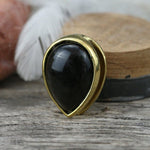 An elegantly designed brooch with a gold frame and golden obsidian stone, much like the Kaya Movement's OBI Minimalist Oval Ear Hangers in Gold & Golden Obsidian, rests on a wooden surface. In the blurred background, a white feather and rustic wooden objects accentuate its sophistication.