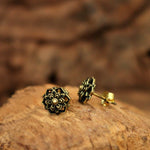 A pair of ILLY Lotus Mandala Stud Earrings in Gold by Kaya Movement, crafted from yellow brass with intricate floral designs, rests on a textured wooden surface. The soft-focused background highlights the detailed craftsmanship of these 18-gauge earrings for your ear lobe.