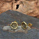 The OM Chakra Stud Earrings in Gold from Kaya Movement rest on a slate stone surface, displaying the Om symbol. The earthy backdrop, featuring shades of brown with a natural texture, enhances their appeal. Crafted from yellow brass with intricate detailing around the edges, these 18 gauge earrings come in elegant packaging for a touch of rustic charm.