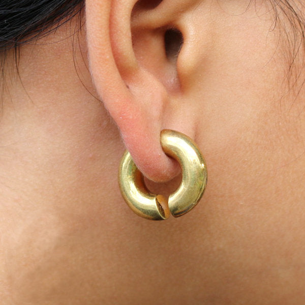 Close-up of a person's ear wearing an ELI Fake Hoop Gauge Earring in Gold by Kaya Movement, featuring a thick, circular design. The 18-gauge earring has a glossy finish and a small gap at the bottom, giving it a slightly open appearance. The image focuses on the ear and earring, with part of the person's hair and skin visible.