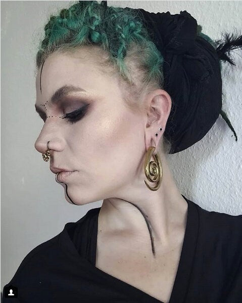 A person with green braided hair adorned with a black headscarf looks to the side. They have dramatic black eye makeup, facial piercings, and unique Brass Spiral heavy ear weights from Kaya Movement in an antique finish. Their outfit is black, and they have a thin black line painted along their jaw and neck.