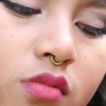 Close-up of a person adorned with the AMA Seamless Nose Ring in Gold (9mm) by Kaya Movement. Their eyes are partially closed, highlighted with black eyeliner, and they are wearing bold, vibrant pink lipstick that accentuates their lips. The image captures a sense of calm and introspection.