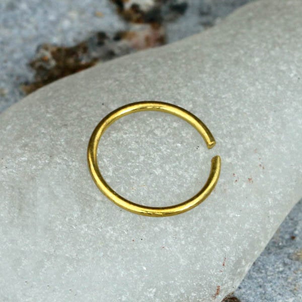 The INDUS Seamless Hoop Ring in Gold by Kaya Movement, a small and simple open-ended gold brass body jewelry ring, is placed on a textured white and gray surface. Its minimalistic design features a smooth, circular shape with a slight gap, giving it an elegant and modern look suitable for multi-use piercing with seamless closure. This ring is 20 gauge in size.
