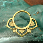 A TWIL Lotus Mandala Septum Ring in Gold by Kaya Movement rests on a blue-green, textured surface. The 18 gauge septum ring features cut-out heart shapes and geometric designs, giving it an ornate and handcrafted appearance.