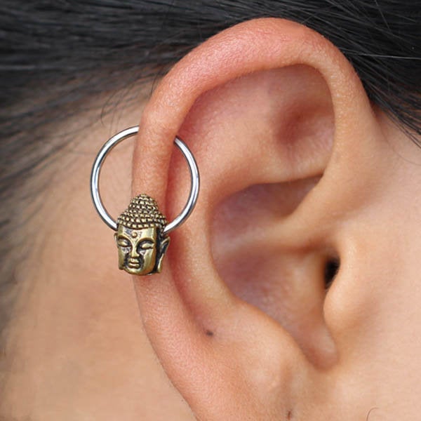 A close-up of a person's ear adorned with the Kaya Movement BUDDHA Silver Ring with Gold Pendant. The multi-use piercing hoop earrings, available in 18, 16, or 14 gauge, feature a small, detailed gold-colored Buddha head charm hanging from them. The person's dark hair is pulled back, accentuating the unique Buddha pendant design.