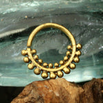 The ELE Seamless Ring in Gold | 18 gauge by Kaya Movement features a small, ornate design with beaded tribal ethnic patterns and tiny spherical decorations around the lower half. The ring is elegantly set against a textured background that resembles wavy glass or water patterns and rests on a wooden surface at the bottom of the image.