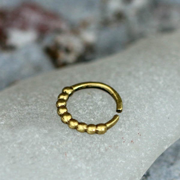 The SOA Single Beaded Ring in Gold from Kaya Movement, a small circular piece of jewelry, rests on a grayish marble surface. This gold brass body jewelry boasts a partially open design featuring one side adorned with a row of small, rounded beads that exude a tribal ethnic design aesthetic.