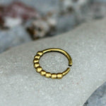 The SOA Single Beaded Ring in Gold from Kaya Movement, a small circular piece of jewelry, rests on a grayish marble surface. This gold brass body jewelry boasts a partially open design featuring one side adorned with a row of small, rounded beads that exude a tribal ethnic design aesthetic.