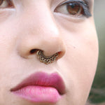 Close-up of a person with winged eyeliner and pink lipstick. They are wearing the AIA Seamless Septum Ring in Gold, an 18 gauge decorative piece from Kaya Movement adorned with small ornate designs, featuring a tribal ethnic flower design. The background is blurred.