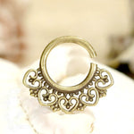 The AIA Seamless Septum Ring in Gold by Kaya Movement is an exquisite piece of jewelry, featuring a delicate, intricate pattern of connected hearts and tribal ethnic flower details. Crafted from yellow brass, this partially open 18 gauge ring hints at its versatility for various piercings such as a nose ring or earring. It is beautifully showcased against a soft, blurred background.