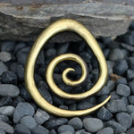 The Kaya Movement Brass Spiral heavy ear weight, an 8mm piece of tribal style jewelry crafted from nickel-free brass with an antique finish, is placed on a surface covered with smooth, dark pebbles. The background shows a section of a stone wall.