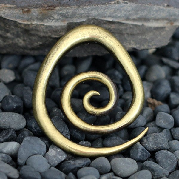 The Kaya Movement Brass Spiral heavy ear weight, an 8mm piece of tribal style jewelry crafted from nickel-free brass with an antique finish, is placed on a surface covered with smooth, dark pebbles. The background shows a section of a stone wall.