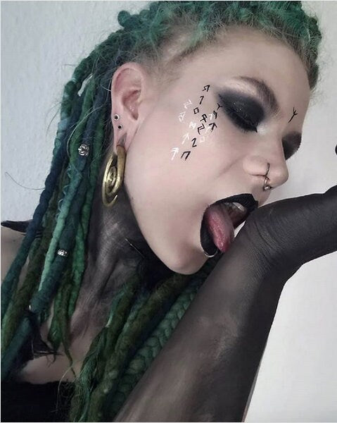A person with green dreadlocks and black eye makeup, face tattoos, and black lipstick holds up their right arm with a black sleeve, appearing to lick it. They have piercings in their ear, nose, and lip, wearing an ornate Brass Spiral heavy ear weight from Kaya Movement. The 8mm gold earring features antique finishing and weighs 42 grams, perfectly complementing their tribal style jewelry.