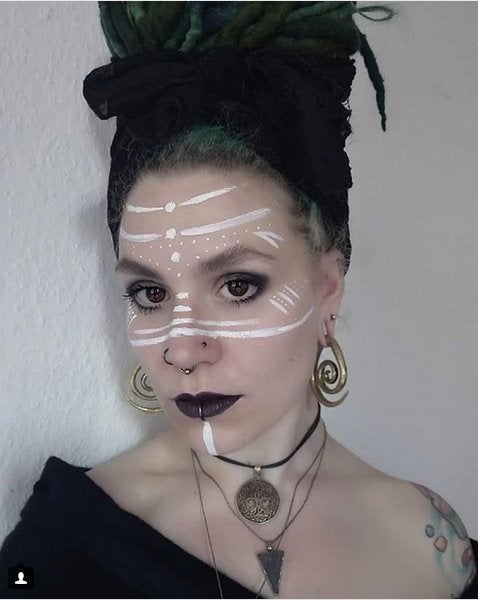 A person with long braided green and black hair wrapped in a headscarf, wearing the SPIRAL Minimalist Ear Hangers in Silver by Kaya Movement, along with necklaces and facial markings in white paint. They have dark lipstick, light skin, and tattooed shoulders adorned with Spiral symbols. The background is plain and light-colored.