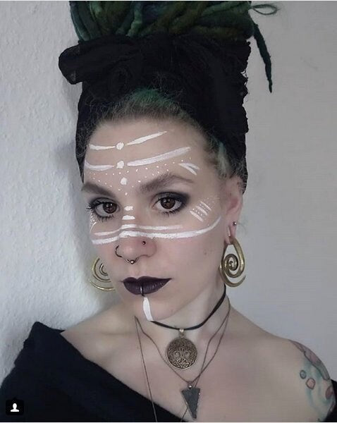 A woman with dark lipstick and face paint designs stands against a plain background. She has a dark headscarf, green and black dreadlocks, large Brass Spiral heavy ear weight earrings (Spirale d'oreille en laiton 8mm, bijoux de Lobe tendu, Ecarteurs Lourds) from Kaya Movement in an antique finish, multiple necklaces made of nickel-free brass in tribal style, and visible tattoos on her shoulder.