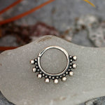 A close-up shot of the BRAI Seamless Septum Ring in Silver by Kaya Movement, made from 925 silver, resting on a light-colored, smooth stone surface. The 18 gauge ring features a beaded design with small, evenly spaced silver beads adorning the bottom half and a touch of tribal ethnic design, giving it an ornate appearance.