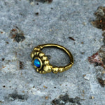 The SHA Beaded Flower Nose Ring in Gold & Black Onyx by Kaya Movement, a 20-gauge piece featuring a central black onyx gem encircled by intricate gold beading in a tribal ethnic style, is showcased on a textured, slightly rough gray surface with hints of brown and red.