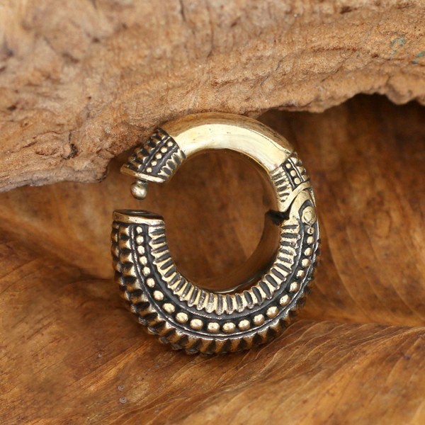 A close-up photograph of a vintage-style, gold earring with intricate detailing. The MAHAL Medium Hoop Ear Weights in Gold with Clicker from Kaya Movement feature a textured pattern and are set against a wooden background. The overall aesthetic is rustic and reminiscent of antique-style brass jewelry.