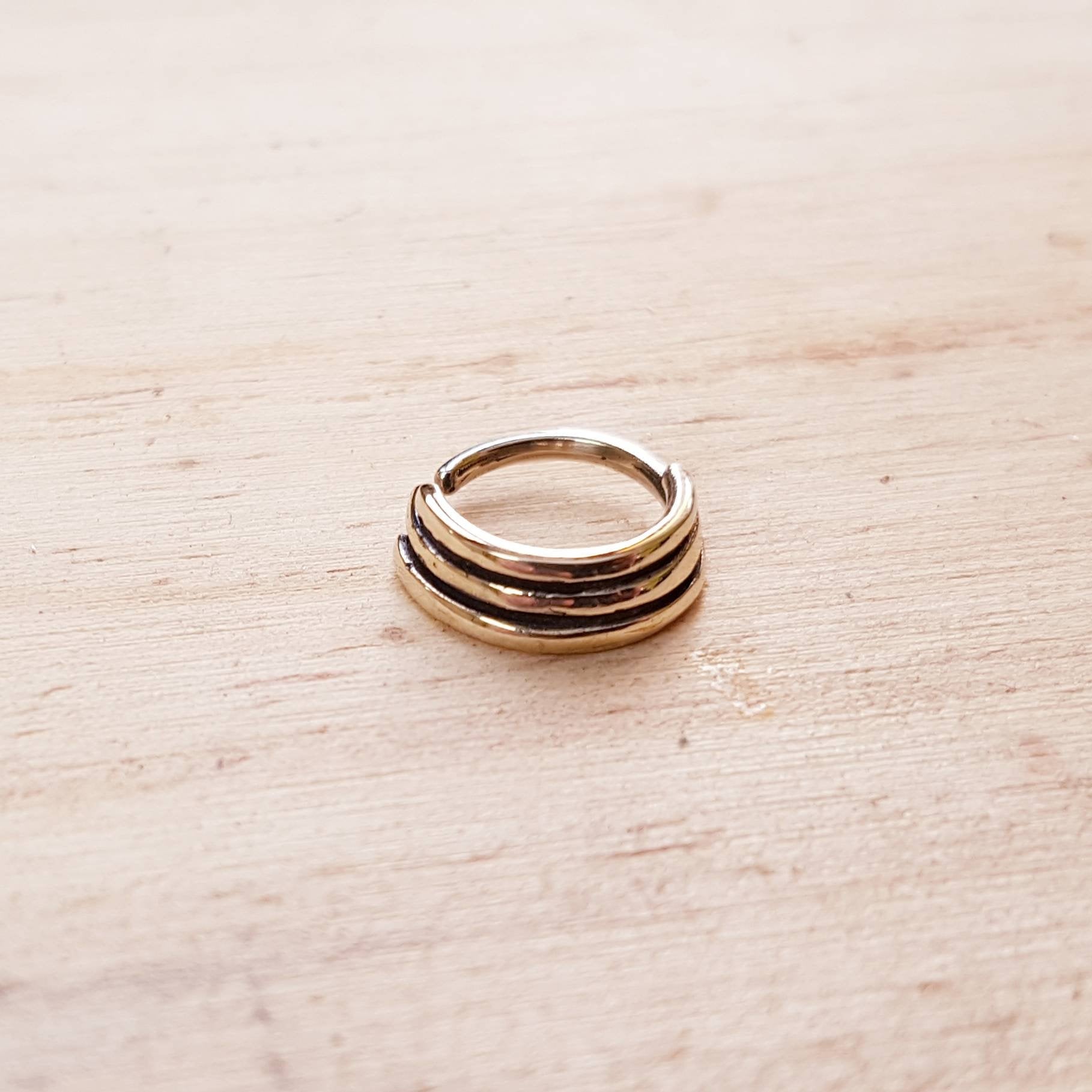 The TRIA Septum Ring in Gold by Kaya Movement, an 18-gauge ring with a minimalist, multi-banded design of three thin stacked bands, rests on a light wooden surface. Its polished finish and sleek, modern appearance evoke the elegance of brass body jewelry.