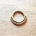 A close-up of the Kaya Movement's IRI Seamless Septum Ring in Gold (16 gauge), adorned with a row of small, evenly spaced beads along the bottom curve, placed on a textured wooden surface, showcasing its tribal ethnic design body jewelry aesthetic.