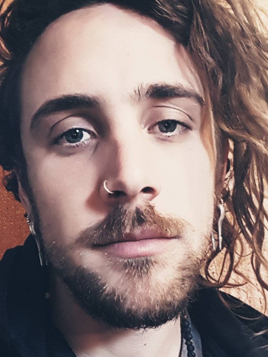 Close-up of a person with light skin and light-colored eyes, their long, wavy hair cascading over one side of their face. They sport a mustache and beard, complemented by 925 silver body jewelry from Kaya Movement, including the INDUS Seamless Minimalist Hoop Ring in Silver (20 gauge) as a nose ring and minimalist earrings. Their expression is neutral.
