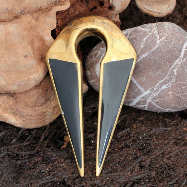 The Brass Ear Weight by Kaya Movement, featuring heavy futuristic black inlay and brass tunnels with a distinct 12mm design, is showcased against a backdrop of natural rock and soil. Reminiscent of piercing body jewelry, this unique piece has a modern, sleek aesthetic.