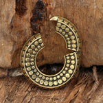 A pair of SILO Seamless Hoop Ear Weights in Gold with Clicker by Kaya Movement sits against a rough wooden background. The ear weights, featuring an intricate textured pattern of raised dots, have a small gap at the top. Their design exudes a rustic, antique feel reminiscent of tribal gold ear weights.