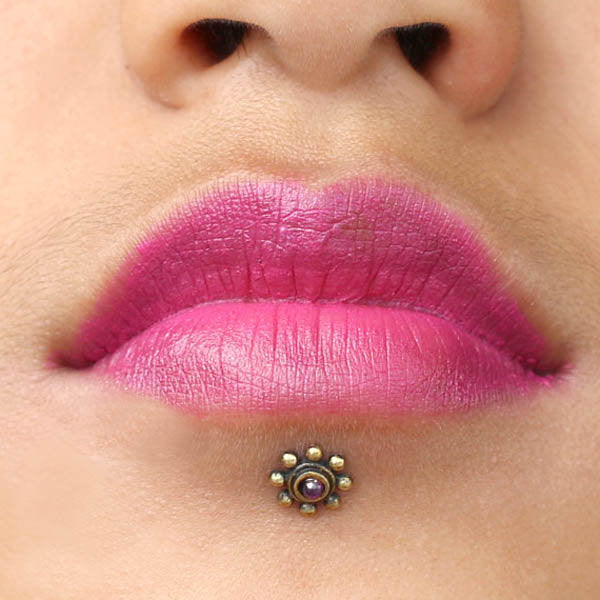 Close-up of a person's lips adorned with bright pink lipstick. Below the lips is a chin piercing featuring Kaya Movement's BEA Flower Labret Stud in Gold & Amethyst, designed with a tribal beaded flower and a natural Amethyst stone at its center. The person's skin tone complements the vivid lip color and the stunning piercing.