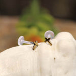 CROSS Labret Studs in Gold from Kaya Movement rest on a white seashell, their clear plastic backs contrasted against a softly blurred background of green and orange hues.