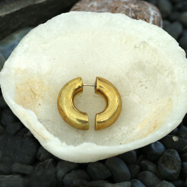 An ELI Fake Hoop Gauge Earring in Gold with a 925 Silver Post by Kaya Movement is displayed on a white, bowl-like shell. The shell, cradling the elegant piece of yellow brass jewelry, rests on a bed of dark, smooth pebbles.