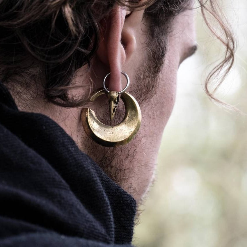 Close-up of a person wearing HOLO Hoop Dangle Ear Weights in Gold, from Kaya Movement, attached to large crescent-shaped brass earrings. The person has long, wavy brown hair and is dressed in a black garment. The background is blurred and depicts an outdoor setting.