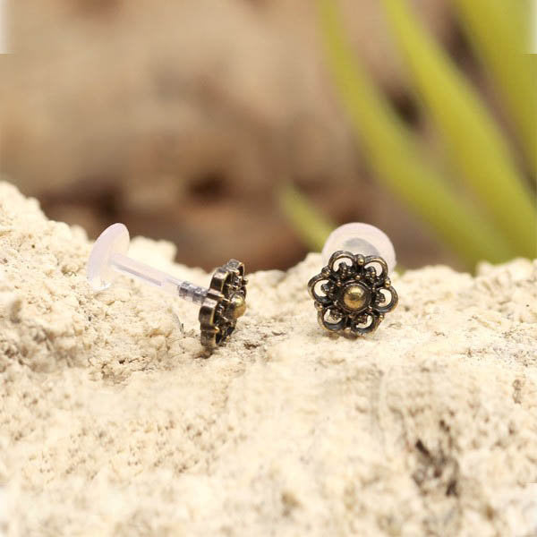 A pair of BLOSSOM Flower Labret Studs in Gold by Kaya Movement, designed with intricate tribal beaded flower patterns, rests on a rough, sandy surface. These vintage-style body jewelry studs feature a small transparent backing and a central gold brass bead surrounded by detailed black metalwork forming the petals.