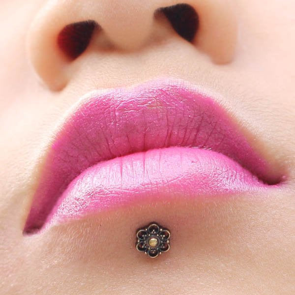 Close-up of a person's lips with bright pink lipstick. The person has a small, BLOSSOM Flower Labret Stud in Gold by Kaya Movement below the center of their bottom lip. The image focuses on the lips and labret stud, with no other facial features visible.