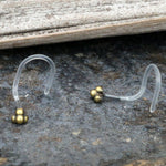 The TRINITY Nose Screw Studs in Gold or Silver by Kaya Movement, featuring clear, curved designs with small clusters of gold-colored beads at the ends, rest elegantly on a textured surface. A wooden plank is partially visible in the background, enhancing the rustic charm of this 20 gauge body jewelry.