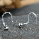 Two transparent TRINITY Nose Screw Studs by Kaya Movement, featuring small, clustered metallic beads at one end, are displayed on a dark, textured surface. A blurred background with earthy tones adds contrast to the clear body jewelry.