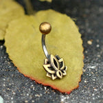 The Lotus Flower Belly Piercing Ring in Gold by Kaya Movement, a 14 gauge navel jewelry piece crafted from surgical steel and featuring a round top ball with a lotus flower design at the bottom, rests on a green leaf with a reddish edge. The leaf is placed on a dark gravel surface. This exquisite ring is perfect for belly button jewelry enthusiasts and comes packaged in beautiful gift wrapping.