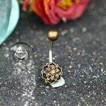 A beautifully designed Lotus Flower Belly Piercing Ring in Gold, 14 gauge, from Kaya Movement is displayed on a textured dark surface. The top ball has a plain gold finish, while the bottom showcases an intricate lotus flower design. In the background, there are blurred flowers and a logo that reads "Kaya Boutique." Gift packaging options are available too.