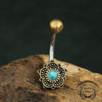 A BLUE LOTUS Flower Belly Piercing Ring in Gold with a natural Turquoise stone, from Kaya Movement, featuring an intricate floral design. The 14 ga surgical steel jewelry rests on a rough surface with a dark background, and the name "Kaya Movement" appears in the lower right corner.
