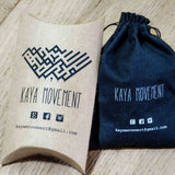 Two packages from Kaya Movement are displayed on a wooden surface. On the left is a cardboard pillow box featuring the company's logo, name, and contact information. On the right is a black drawstring pouch with similar branding and details, making it an ideal gift for their exquisite handmade brass Lotus Flower Pair earrings.