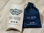 Two packages from Kaya Movement are displayed on a wooden surface. On the left is a cardboard pillow box featuring the company's logo, name, and contact information. On the right is a black drawstring pouch with similar branding and details, making it an ideal gift for their exquisite handmade brass Lotus Flower Pair earrings.