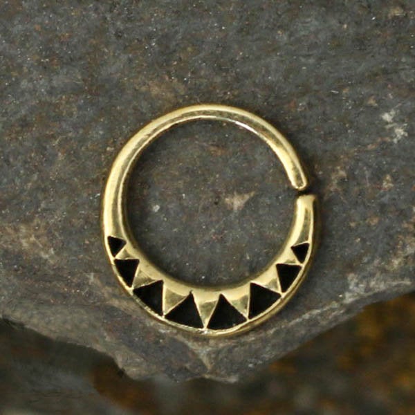 The AYM Seamless Septum Ring in Gold by Kaya Movement is a circular brass body jewelry piece featuring a gold-colored finish. Its semi-circle design showcases small black triangular accents at the bottom, beautifully set against a textured stone background. The seamless ring, available in 18 gauge, includes a small opening at the top for easy insertion and is perfect for various piercing options.