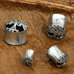 Four Sacred Tree of Life Round Saddle Tunnels in Silver by Kaya Movement, varying from 8mm to 16mm gauge, displayed on a textured brown surface resembling a leaf. These ear gauges feature intricate tree of life designs and resemble brass double flared plugs, with two larger sizes and two smaller sizes.