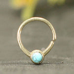 The NAE Seamless Septum Ring in Gold & Turquoise by Kaya Movement, an 18 gauge minimalist piece in gold brass, features a small and round design with an open end showcasing a natural blue turquoise stone at the bottom center, all set against a blurred background.