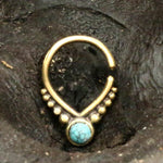 A close-up image shows an 18g Unique Gold Triangle Septum Ring with an open end. The ring, from the Kaya Movement brand, is adorned with an opal stone at the bottom center, surrounded by gold beading on either side. It is placed on a rough, dark textured surface.