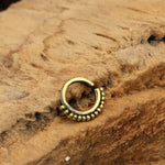 A small IRI Seamless Tragus Ring in Gold (18 gauge) from Kaya Movement sits on a rough, textured wooden surface. The body jewelry is positioned in a slight arc, highlighting its circular shape and intricate tribal ethnic design at the bottom.