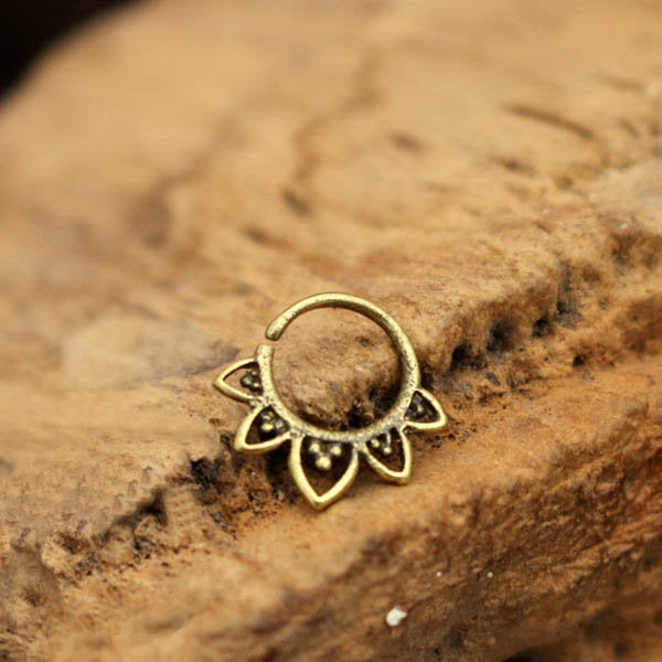 A KAAL Lotus Ring in Gold, meticulously crafted by Kaya Movement, rests on a textured wooden surface. This 20 gauge body jewelry features delicate petal-like shapes that are evenly spaced around its circular form, showcasing an intricate floral pattern with a hint of tribal ethnic design.