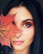 A woman with dark hair and a nose ring holds a large autumn leaf partially covering her face. Her visible eye, adorned with makeup, glints like a black onyx gemstone as she wears the Kaya Movement SYN Seamless Septum Ring in Gold & Onyx | 18 gauge. She has a focused, thoughtful expression against the dark background that highlights her and the vibrant leaf.