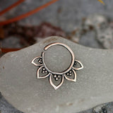 A 925 silver body jewelry piece with intricate detailing sits on a frosted surface. The MAWAR Lotus Flower Septum Ring in Silver by Kaya Movement, an 18-gauge septum nose daith rook helix ring, is adorned with small, petal-like shapes featuring dot patterns inside each petal that give it a floral appearance. The background includes slightly blurred natural elements.